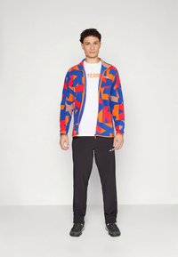 Casaco colorido com fecho éclair e formas abstratas em azul, vermelho e laranja, usado sobre uma t-shirt branca. Calças pretas e sapatos desportivos completam o conjunto.