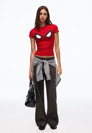 Ung kvinde iført en rød Spider-Man t-shirt, mørke vidde bukser, ternet skjorte bundet om livet, holder en sort håndtaske.