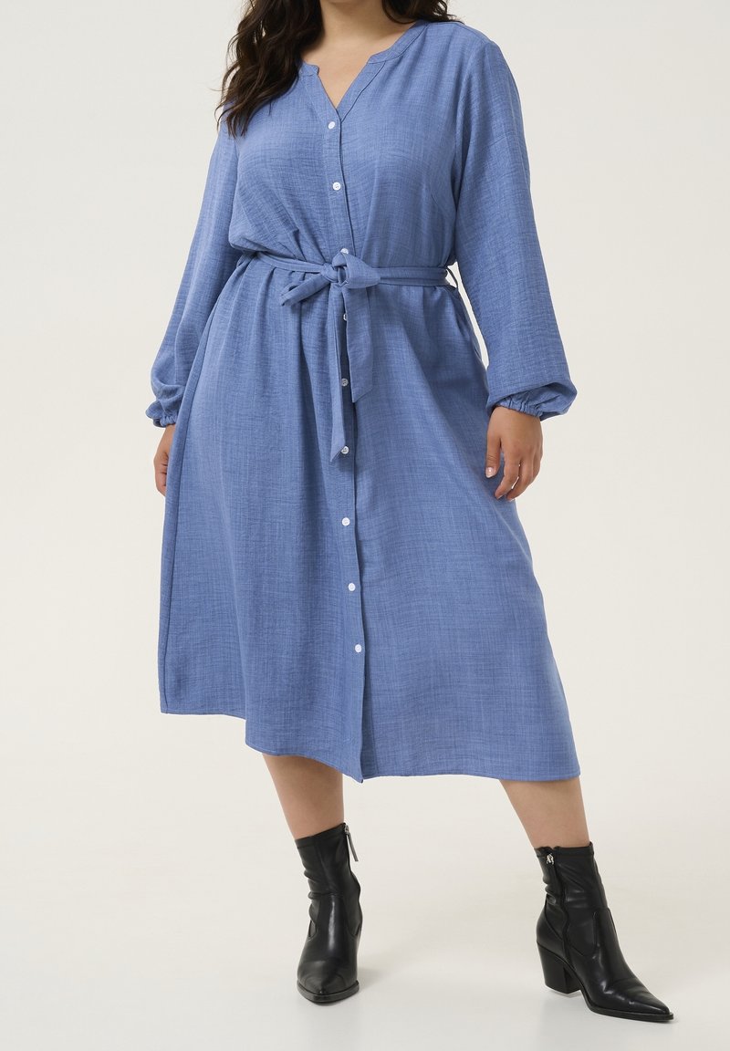 Femme portant une robe bleue à manches longues, boutonnée, avec ceinture à la taille et bottines noires sur un fond uni.