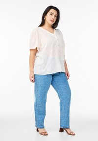 Blusa button-up di un beige chiaro con maniche a sbuffo corte, tessuto testurizzato. Abbinata a jeans blu ad alta vita con texture e sandali con tacco marroni.