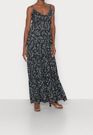 Kobieta w czarnej, kwiatowej maxi sukience na cienkich ramiączkach z falbanami, w brązowych sandałach na obcasie z paskami, stojąca na szarej podłodze.