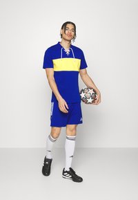 Conjunto atlético azul y amarillo que incluye una camiseta de manga corta con una franja amarilla horizontal y pantalones cortos a juego, combinados con botas de fútbol negras.