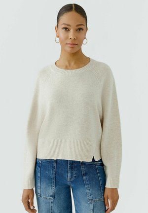 Jonge vrouw draagt een crèmekleurige gebreide trui met geribde zoom en manchetten, gecombineerd met blauwe jeans met grote voorzakken.