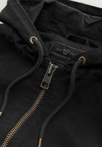 Gros plan d'une veste à capuche noire avec une fermeture éclair métallique, des cordons noirs avec embouts métalliques et une étiquette en tissu à l'intérieur du col.