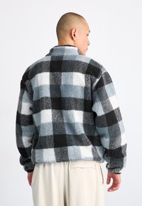 Fleece pulóver fekete, szürke és fehér kockás mintával, magas gallérral és textúrált anyaggal, laza szabású és ejtett vállú.