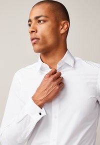 Hvid skjorte med knapper og spids krave, med sorte detaljer på manchetterne. Glat tekstur og skræddersyet pasform.