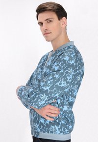 Giacca bomber blu oversize con motivo floreale in diverse sfumature, dotata di colletto e polsini a coste e chiusura zip.