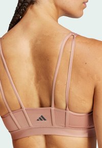 Sujeción deportiva de color rosa claro con tirantes finos, que presenta un acabado texturizado, suavidad elástica y un pequeño logo negro en la parte posterior.