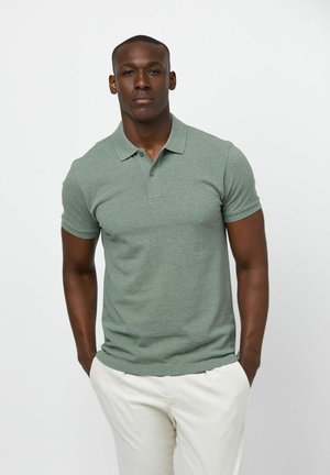 Camiseta tipo polo de manga corta en algodón verde apagado, con cuello clásico, tapeta de tres botones y diseño ajustado. Combinada con pantalones de color claro.