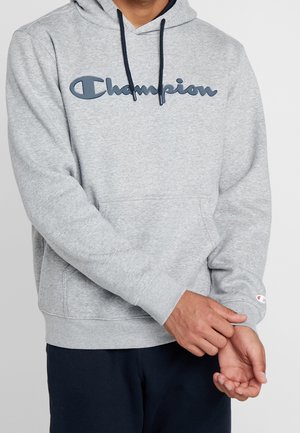 Persoon draagt een lichtgrijze Champion-hoodie met marineblauwe trekkoorden, een blauw logo op de borst en een klein logo op de linker pols en past de mouw aan.
