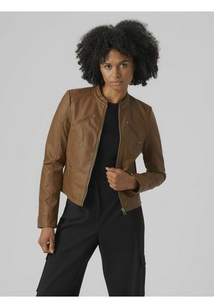 Veste en cuir marron au design court, avec col montant, fermeture zippée et coutures diagonales. Portée par-dessus un haut noir et un pantalon.
