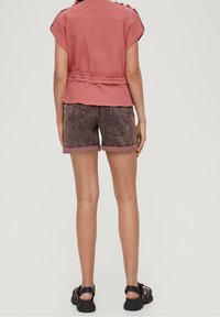 Femme portant un haut court à manches courtes en rose poussiéreux texturé et un short en denim foncé retroussé, avec des sandales noires, se tenant de dos.