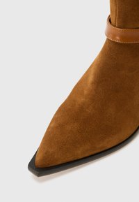 Botte chelsea en daim marron avec un bout pointu, dotée d'une sangle de cheville en cuir marron et d'une semelle en caoutchouc noir. La texture lisse met en valeur le design.