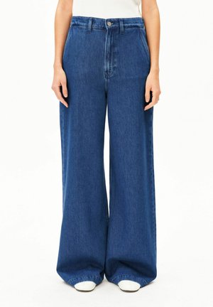 Personne portant un jean bleu taille haute à jambes larges et un haut blanc, debout les bras détendus le long du corps devant un fond blanc uni.