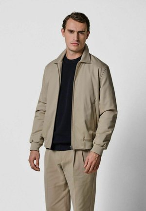 Beige leichte Jacke mit Stehkragen und durchgehendem Reißverschluss, kombiniert mit einem dunkelblauen Pullover und passenden Hosen, ausgestattet mit gerippten Bündchen.