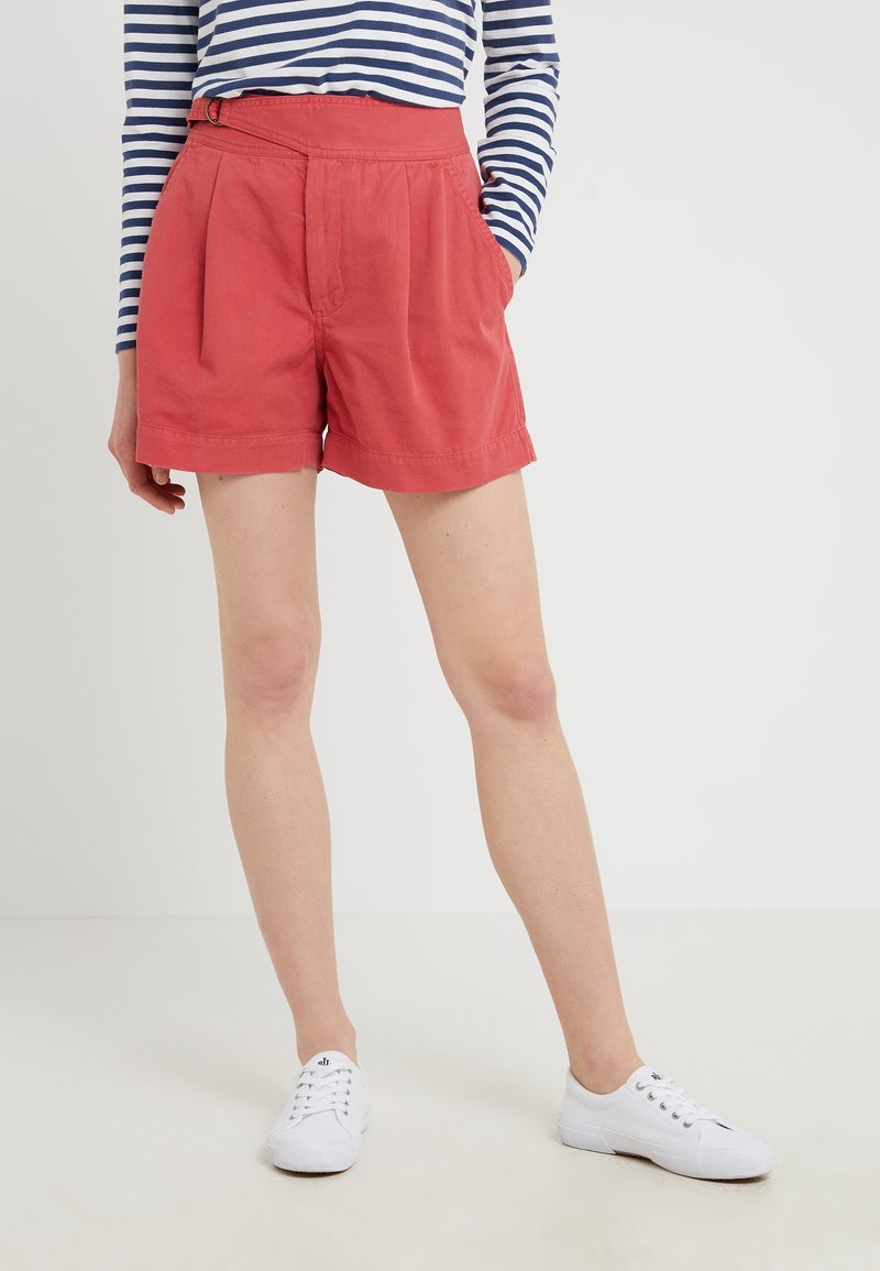 Pantaloncini rossi in cotone con pieghe e tasche laterali, dotati di dettaglio in vita. Abbinati a una maglietta a maniche lunghe a righe navy e bianche e a sneakers bianche.