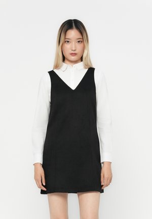 Rochie neagră fără mâneci, cu decolteu în V, suprapusă peste o cămașă albă cu guler. Design simplu, textură netedă, se termină deasupra genunchiului.