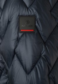 Detail tmavě modré prošívané péřové bundy s texturovanou látkou, diagonálním kosočtverečkovým vzorem a černou nášivkou s červeným akcentem.