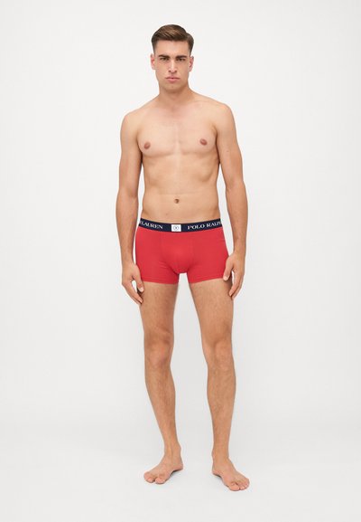 Crvene bokserice s mornarskoplavim pojasom s natpisom "POLO RALPH LAUREN." Tkanina se čini glatkom i rastezljivom, čvrsto prianjajući uz tijelo.