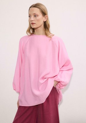 Vaaleanruskeatukkainen nainen, joka pukeutunut väljään vaaleanpunaiseen pitkähihainen yläosaan ja tummanpunaiseen housuihin, seisoo käsi taskussa ja katsoo vasemmalle.