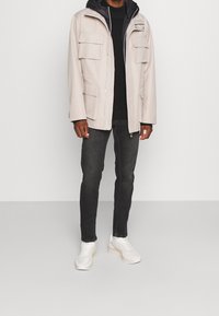 Ljusgrå jacka med fyra framfickor, dragkedja framtill och huva; bärs över en svart tröja, matchad med mörka jeans och vita sneakers.