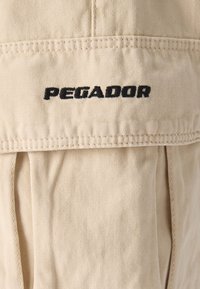 Béžová tkanina s texturovaným povrchem, s prošitými detaily; logo "PEGADOR" v černé výšivce na klopě kapsy.