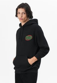 Black hoodie made of soft fabric featuring a front pocket. Displays "HUF" logo in green and red on the left chest area.