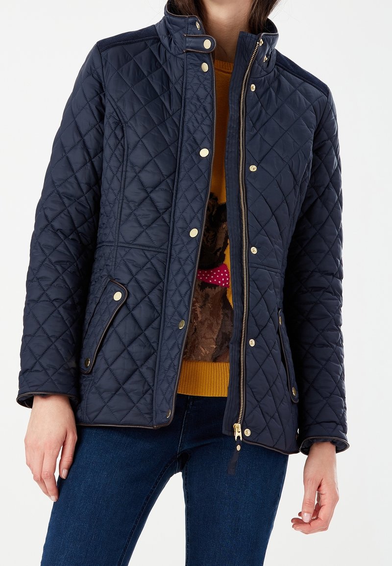 Femme portant une veste matelassée bleu marine avec des boutons dorés et une fermeture éclair, sur un pull moutarde avec un chien orné d'un nœud rose à pois.
