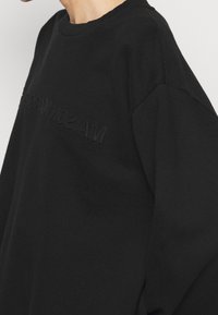 Černá mikina má design s kulatým výstřihem a jemně tónované logo vyšívané na přední straně, což jí dodává uvolněný, oversize střih.