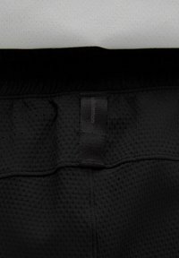 Crne atletske šortse s teksturiranim materijalom, s čvrstim pojasom i oznakom od materijala sprijeda. Bijela pozadina s malim perforacijama.