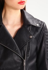 Kvinna klädd i svart läderjacka med dragkedja och tryckknappar, röd läppstift synligt på hennes något vända ansikte, enkel ljus bakgrund.