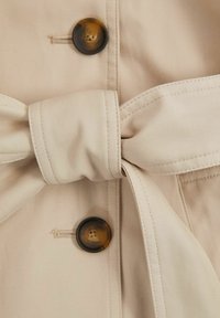 Béžová trench coat tkanina s velkým tmavě hnědým knoflíkem a zavázaným páskem. Texturováno s viditelnými švy.