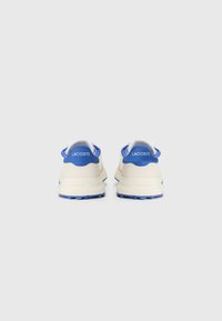 Lacoste sneakers in cream with blue accents. Features a smooth upper, padded collar, and textured sole for grip. Logo on heel.