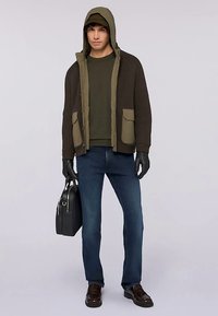 Tmavě zelená a hnědá žebrovaná bunda se dvěma kapsami, přehozená přes zelenou košili, doplněná modrými džínami a černými rukavicemi. Součástí je černá taška a čepice.