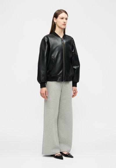 Žena stojaca, oblečená v čiernej koženej bomber bunde, svetlosivých širokých nohaviciach a čiernych špicatých balerínach, na jednoliatom svetlom pozadí.