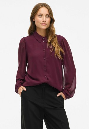 Burgundy button-up blouse with long sheer sleeves, puffed shoulders, and a classic collar. Paired with black trousers. Smooth texture.