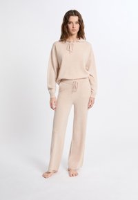 Set di abbigliamento da casa di colore beige chiaro realizzato in materiale morbido. Include un maglione con cappuccio e coulisse e pantaloni larghi con vita elasticizzata.