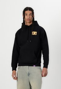 Svart huvtröja med framficka. Har orange grafisk text "JACKER STREET FOOD." Avslappnad passform, mjuk textur, kombinerad med ljusa jeansbyxor.