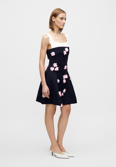 Abito blu navy con corpetto aderente, scollo arricciato bianco e applicazioni floreali rosa, gonna svasata, abbinato a mules bianche con tacco.