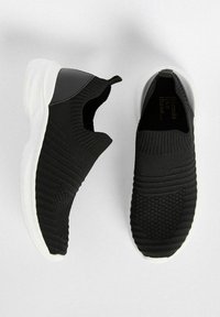 Černé slip-on tenisky s texturovaným pleteným svrškem, které mají černý akcent a bílou gumovou podrážku. Podrobnosti pleteného vzoru po stranách.