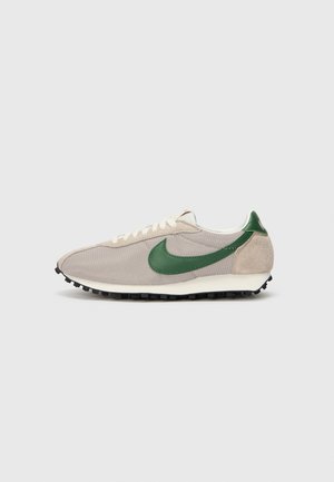 Beige løbesko med en tekstureret mesh-overdel, grønne ruskindsdetaljer, hvide snørebånd og en sort gummisål. Har et klassisk swoosh-design.