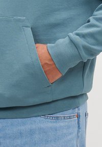 Sweatshirt azul claro com um grande bolso frontal, punhos e cós canelados. Textura suave, design casual. Usado sobre jeans de denim claro.