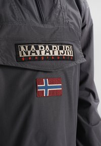 Grå anorak med en forlommetask, der har et sort broderet mærkelogo og et rødt mærke med et blåt og hvidt flagdesign.