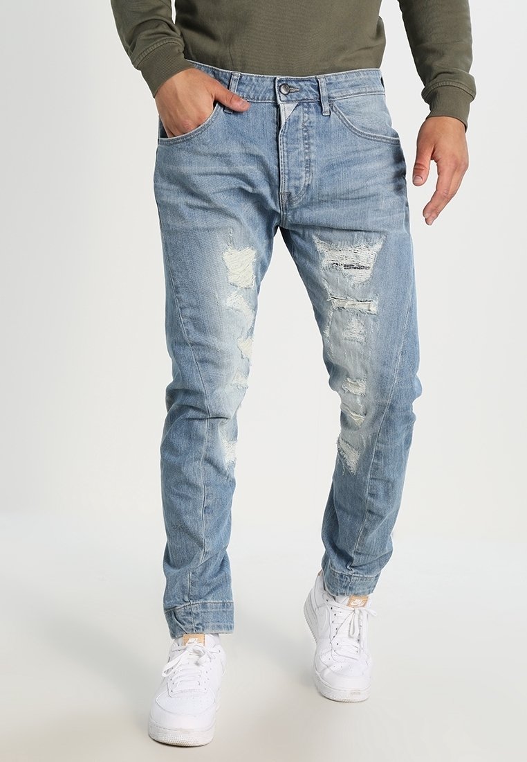 Homme portant un jean bleu clair déchiré, une main dans la poche, assorti à des baskets blanches et un haut à manches longues vert olive.