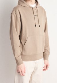 Sudadera beige con un bolsillo frontal, capucha con cordón y sutil detalle del logo en negro. Tejido suave con puños y cinturilla acanalados.