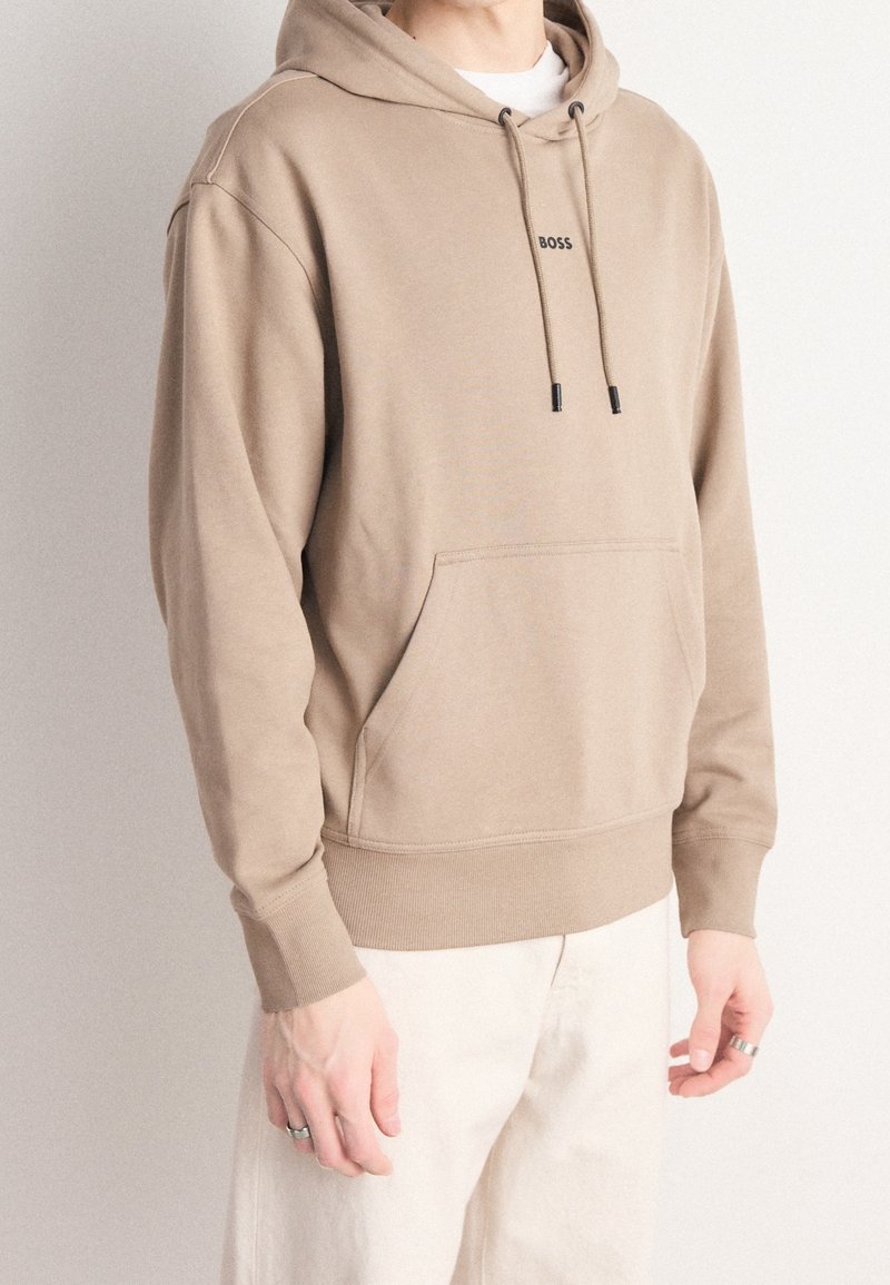 Sudadera beige con un bolsillo frontal, capucha con cordón y sutil detalle del logo en negro. Tejido suave con puños y cinturilla acanalados.