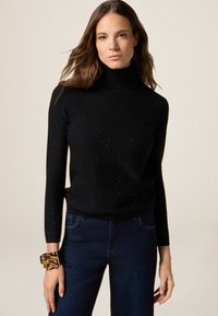 Pull à col roulé noir avec des manches longues, arborant une texture mouchetée. Porté avec un jean en denim foncé et un bracelet métallique au poignet.