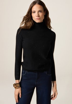 Pull à col roulé noir avec des manches longues, arborant une texture mouchetée. Porté avec un jean en denim foncé et un bracelet métallique au poignet.