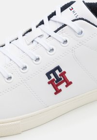 Zapatilla blanca hecha de cuero con un forro negro texturizado, con cordones blancos, ojales de metal y un logotipo bordado en rojo y azul marino.
