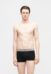 Młody mężczyzna o jasnej karnacji i zaczesanych do tyłu włosach, ubrany w czarne bokserki Calvin Klein, stojący na jednolitym jasnym tle.